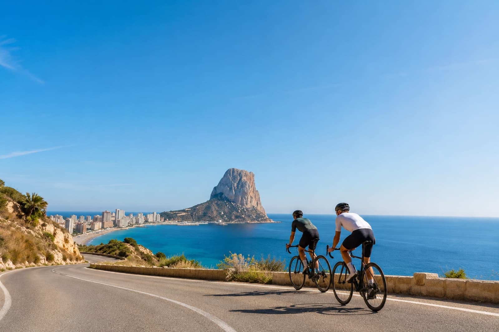 Two road cyclists on the coast road above Calpe bay, with the Peñón de Ifach behind